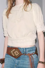 Blusa color crudo de cuello redondo y manga corta con puños. Presenta bordados a tono y cierre trasero con botones forrados.