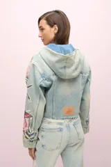 Campera de jean celeste con lavado gastado, corte cropped y capucha de jean. Presenta cierre frontal con botones metálicos, bolsillos delanteros y bordados florales en las mangas.