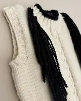 Chaleco tejido a mano color crudo, con cuello y borde de mangas en negro, y flecos negros en los laterales.