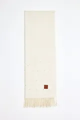 Pashmina color blanco roto con flecos en los extremos y decorada con perlas pequeñas. Incluye un parche de cuero sintético marrón con un logo grabado.