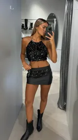 Minifalda de cuero sintético color negro, de tiro bajo, con detalle de cinturones decorativos con ojales metálicos y hebillas en la parte superior.