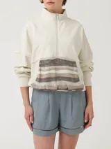 Buzo color beige con cierre frontal parcial y cuello alto. Presenta un bolsillo tipo canguro con diseño tejido a rayas horizontales en tonos marrón, beige y blanco.