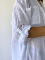 Vestido camisero blanco de corte holgado, con cuello mao, cierre frontal con botones, mangas 3/4 con puños abotonados, dos bolsillos de parche en el pecho y aberturas laterales en el ruedo.