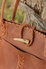 Bolso tipo tote de cuero color marrón con detalles de costuras trenzadas en los bordes y bolsillo externo. Tiene cierre imantado interno y broche con pasador de guampa en el exterior.