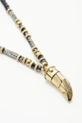Collar largo con cadena compuesta por cuentas de madera pintada en negro y blanco, cuentas metálicas doradas y cuentas ovaladas negras. Dije central metálico dorado con forma de cuerno con texturas.