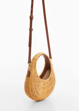 Bolso tipo canasta de mimbre color beige con asa corta circular y correa larga de cuero marrón.