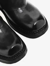 Bota alta de cuero negro con diseño de caña ancha y efecto de solapa que cubre el talón. Presenta detalles de tachas metálicas en la costura posterior, punta redondeada y suela con taco bajo.