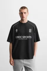 Camiseta negra de corte boxy fit, cuello redondo y manga corta. Presenta logos estampados en blanco en el frente y detalles en verde en los laterales.