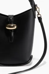 Cartera tipo balde negra de cuero sintético con asa de mano rígida y correa bandolera ajustable y extraíble. Presenta un cierre con trabilla y hebilla metálica dorada.