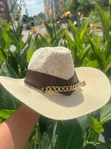 Sombrero de ala ancha tejido en hilo de coco y sintético, color marrón claro. La copa está cubierta con encaje naranja quemado con patrón floral. Tiene una cinta de cuero marrón oscuro y un aplique decorativo de hoja de gamuza con incrustaciones de pedrería.