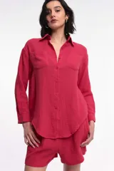 Camisa color fucsia de tela arrugada, con cuello clásico, cierre frontal con botones, dos bolsillos de parche en el pecho y mangas largas con puños abotonados.