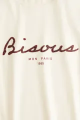 Remera color crema de corte holgado en punto de algodón. Modelo con cuello redondo y estampado frontal con la palabra "Bisous" en color borgoña y la frase "Mon Paris 1985" en negro.