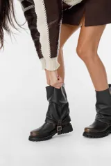 Bota corta de cuero negro con caña arrugada, suela track y correa con hebilla decorativa.