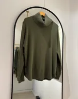 Sweater verde seco de cuello alto envolvente, corte recto y detalle central tejido en espiga. Presenta botones decorativos en los laterales.