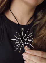 Collar con dije de forma orgánica que simula una estructura de coral o una estrella de mar, hecho en metal plateado con detalles dorados. El dije cuelga de un cordón negro fino con un pasador cilíndrico plateado.