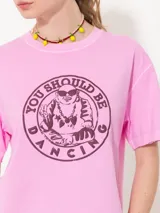 Remera rosa de algodón pima con estampado circular en el frente con la frase 'You Should Be Dancing' y un personaje bailando. Tiene corte amplio, mangas cortas, cuello redondo y tajos laterales.