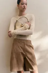 Cartera tipo bucket color beige, con asa de mano tubular y correa cruzada ajustable y extraíble. Cierre con trabilla y adorno metálico dorado en el frente. Forrada.