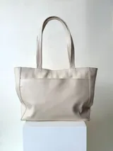 Cartera tipo tote color beige, hecha en cuero graneado con textura uniforme y suave al tacto. Tiene doble asa de hombro, un bolsillo frontal con broche imantado, un bolsillo trasero con cierre metálico y cierre principal también metálico. El interior está forrado y cuenta con tres bolsillos: uno con cierre, uno tipo plaqué para celular y un bolsillo grande tipo sobre con solapa y botón.