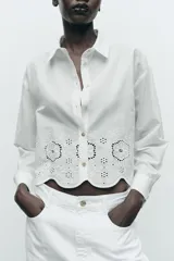 Camisa blanca corta de cuello solapa y manga larga. Bajo con bordados perforados combinados a tono. Cierre frontal con botones.