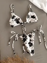 Conjunto de bikini blanco con estampado de flores negras. El corpiño es triangular con tirantes regulables y la bombacha es regulable con tiras en los costados, pudiendo usarse como vedetina o colaless.