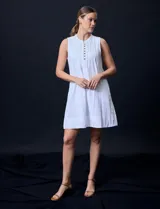 Vestido corto blanco de algodón y rayón, sin mangas, con cuello a la base, abertura central con botones y detalle plisado en la delantera. Incluye bolsillos laterales.