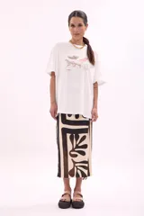 Remera blanca de algodón, corte oversize, con cuello redondo y manga corta. Presenta un estampado frontal con varias reposeras y hamacas de playa, y el texto "LA SIESTA".