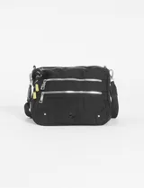 Bandolera negra de tela con múltiples cierres horizontales plateados. Incluye una correa ajustable y detalles colgantes en los tiradores de los cierres, uno de ellos es una borla trenzada blanca y negra y otro es un tirador pequeño de color amarillo.