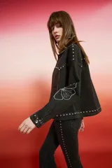 Campera de jean negra de corte holgado con detalles de tachas plateadas en costuras, hombros y puños, y bordado floral blanco en las mangas.