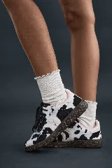 Championes de estilo deportivo con estampado de vaca (manchas negras sobre fondo blanco), cordones negros y suela de goma marrón oscuro. Presentan detalles en rosa pálido en el borde del cuello.