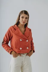 Blazer corto color ladrillo, de paño de lana, con doble botonadura y cuello solapa. Presenta apliques de cuero en codos y puños, y bolsillos de parche.