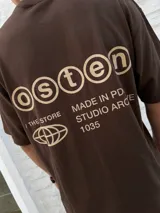 Remera marrón de manga corta, corte oversize, con un pequeño logo de un globo terráqueo estilizado y el texto "STUDIO ARCHIVE" estampado en el pecho.
