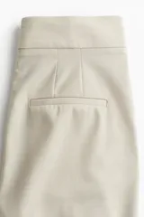 Pantalón cigarette color beige de tiro alto, con cierre y corchetes ocultos, bolsillos laterales discretos y bolsillos insertados decorativos atrás. Piernas ajustadas con abertura en los bajos y pliegues adelante y atrás.