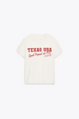 Camiseta color crudo de algodón con efecto lavado, cuello redondo, manga corta con hombro caído y estampado frontal con la leyenda "Texas USA Ranch Original est. 1975" en color rojo.