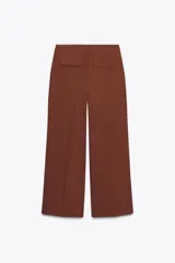 Pantalón marrón de tiro alto con bolsillos delanteros y falsos bolsillos traseros con tapeta. Presenta pliegues en la parte delantera y cierre frontal con cremallera y gancho metálico.