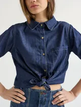 Camisa de denim azul con costuras blancas a contraste, corte crop y cierre frontal con botones. Presenta cuello clásico, manga corta y un bolsillo de parche en el pecho. Se ata en la parte delantera.