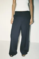 Pantalón negro con finas rayas verticales blancas, de tiro medio y corte ancho. Presenta bolsillos delanteros y traseros, cintura doble tipo bóxer con botones y cierre frontal con cremallera y ganchos metálicos. Detalle de franja marrón en los laterales.