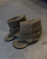 Bota tipo sandalia de gamuza color taupe, con caña arrugada y correa ajustable con hebilla metálica plateada alrededor del tobillo. Diseño tipo chinela con tira entre los dedos.