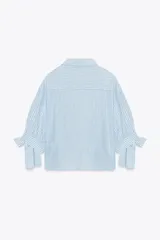 Camisa celeste y blanca a rayas verticales, confeccionada con lino e hilo metalizado. Tiene cuello solapa, mangas 3/4 con lazos en los puños y cierre frontal con botones ocultos por solapa.