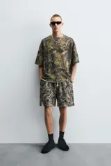 Sudadera relaxed fit con estampado de hojas en tonos verde, marrón y negro. Tiene cuello redondo, manga corta y bajo irregular con aberturas laterales.