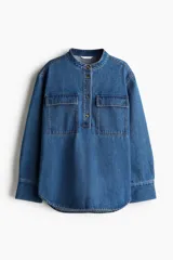 Camisa de denim azul de corte holgado, con cuello mao, tapeta de botones, hombros caídos, mangas largas con puños abotonados y dos bolsillos superiores con solapa.