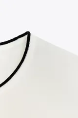 Top blanco de punto con cuello redondo y manga corta. Detalle de ribete negro en cuello, mangas y ruedo.
