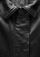 Abrigo negro oversize de cuero ovino con cuello alto y cierre frontal con cremallera y botones ocultos.