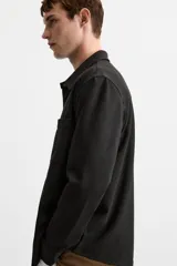 Sobrecamisa negra de tejido con elasticidad, cuello solapa y manga larga acabada en puño con botón a presión. Tiene bolsillos de plastrón en el pecho y cierre frontal con botones a presión.