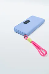 Billetera rectangular color fucsia con cierre perimetral y parche frontal negro con logo. Incluye correa de mano de cordón rosa neón y detalle de ajuste en verde lima.