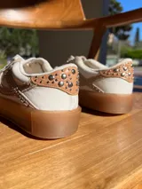 Championes de cuero color beige y blanco, con tachas plateadas en los laterales y suela de goma color caramelo.