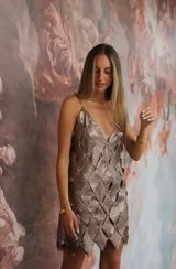 Vestido corto color peltre (marrón grisáceo) con tirantes finos de cadena. El cuerpo del vestido está construido con paneles superpuestos en forma de rombos, unidos por ojales metálicos con forma de flor.