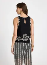 Blusa negra sin mangas con cuello redondo y detalles bordados en contraste color crudo en cuello, sisas y bajo. Presenta apliques ornamentales en el centro de la parte delantera y trasera, detalles calados con transparencias y abertura en forma de lágrima en la espalda con cierre de un botón.