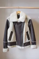 Campera gamulán marrón con interior de piel sintética blanca, cierre frontal con cremallera, cuello con solapas y dos bolsillos con cierre.