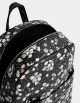 Mochila negra con estampado de margaritas blancas, confeccionada en cuero sintético. Cuenta con correas regulables, manija superior, acceso con cierre, bolsillo frontal con cierre y tag de cuerina. Incluye un pouch extraíble.