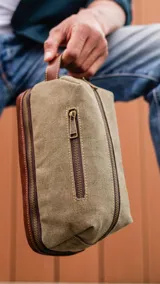 Neceser verde militar de tela canvas con detalles en cuero marrón. Tiene un bolsillo frontal con cierre, asa lateral y doble compartimento con cierre en la parte superior y en la base.